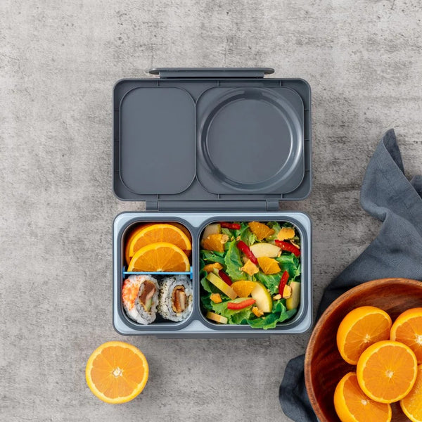 Lunchbox mit Thermosbehälter OmieBox Up – Graphite gefüllt mit frischem Obst, Sushi und Salat auf grauem Untergrund