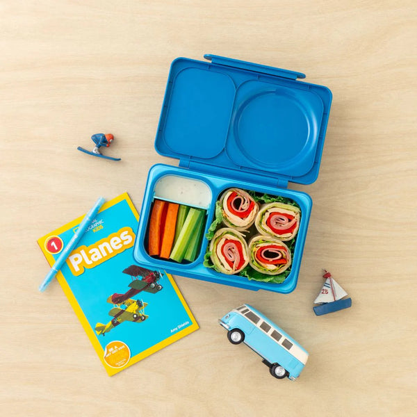 Lunchbox mit Thermosbehälter OmieBox Up – Cosmic Blue gefüllt mit Gemüse und Wraps neben Spielzeug und Buch