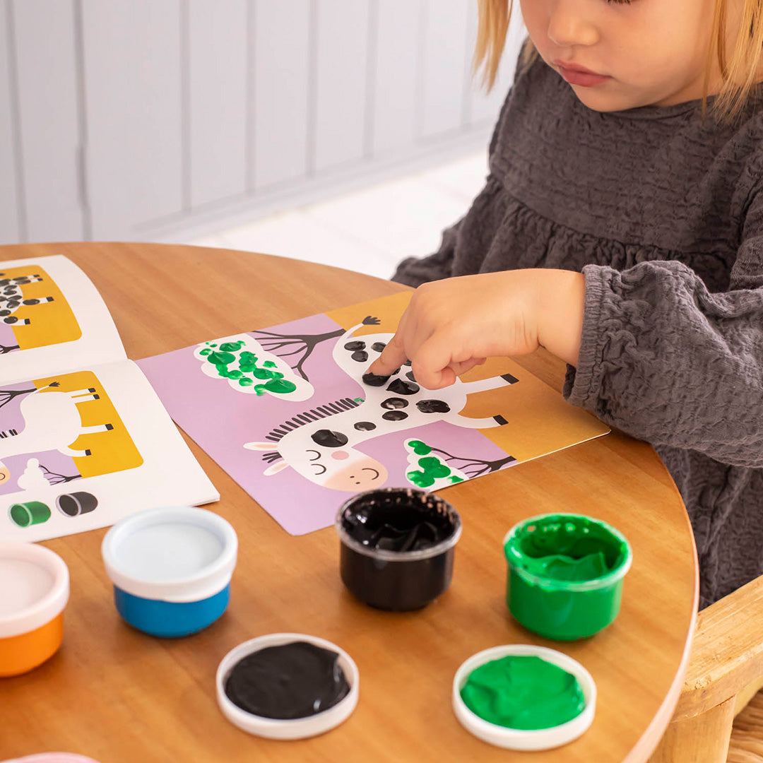 Kind malt mit Fingerfarben ein Bastelbild mit Tieren auf einem runden Holztisch Spielzeug zum Basteln