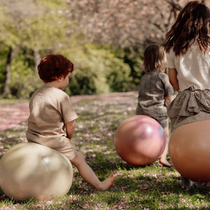 Kinder und Erwachsene spielen draußen mit Hüpfball Grün auf einer Wiese unter blühenden Bäumen