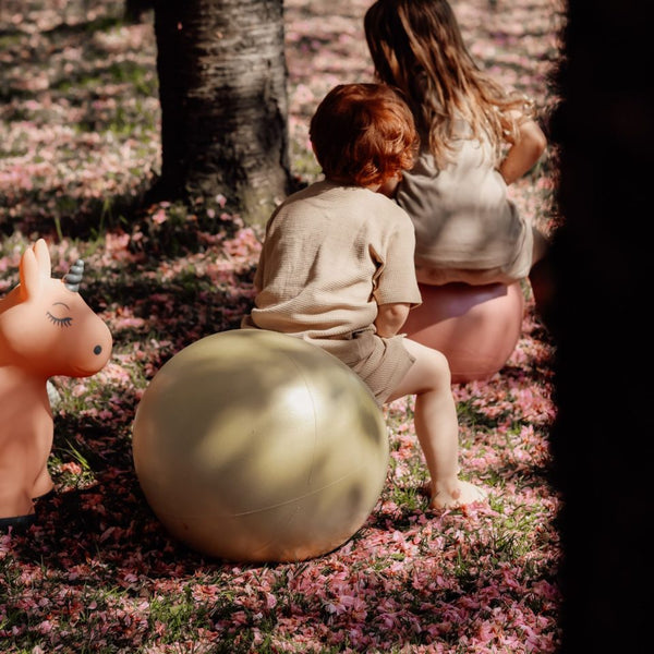 Kinder spielen draußen auf Hüpfball Grün und einem pinken Hüpfball auf mit rosa Blüten bedecktem Gras