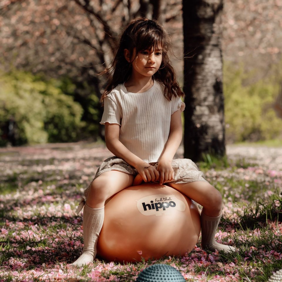 Mädchen sitzt draußen auf einem orangefarbenen Hüpfball Orange von Find that Hippo im Park