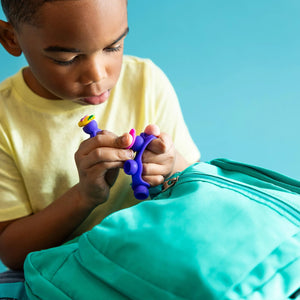 Kind spielt mit dem Schmuckset Plip Charms – Violett von Fat Brain Toys vor einem türkisfarbenen Rucksack