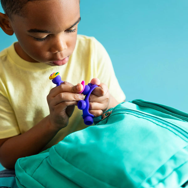 Kind spielt mit dem Schmuckset Plip Charms – Violett von Fat Brain Toys vor einem türkisfarbenen Rucksack