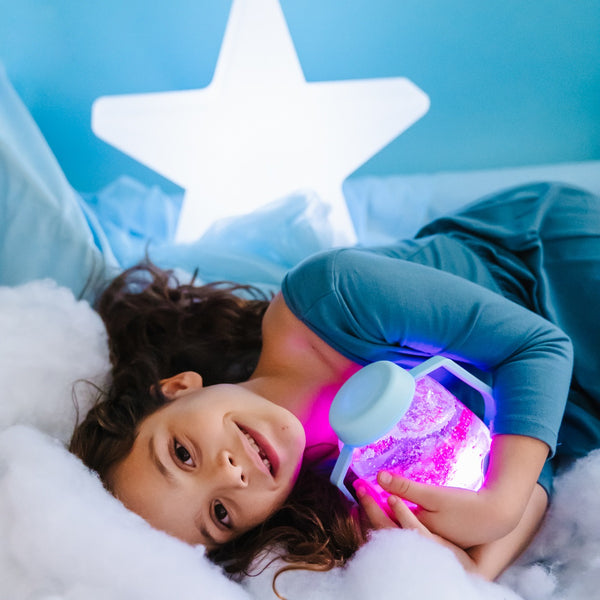 Kind mit Glo Pals Zabawka progresywna Sensorik-Glas zum Befüllen mit Wasser – hellblau liegend im Bett mit leuchtendem Glas