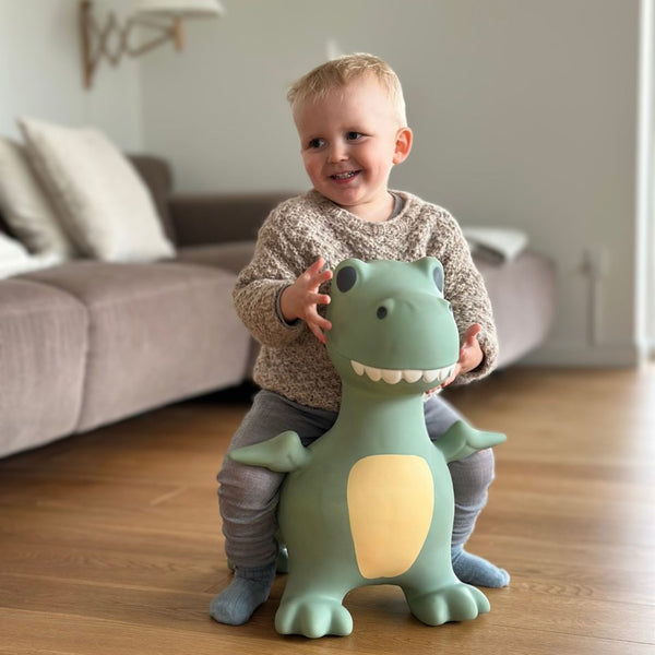 Kleines Kind sitzt freudig auf dem grünen Springer Dinosaurier von Find that Hippo im Wohnzimmer