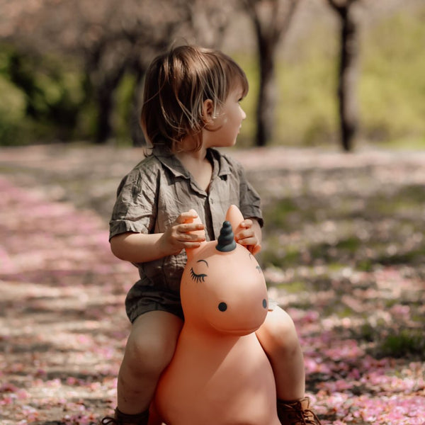 Kind sitzt lachend auf einem orangefarbenen Springer Einhorn Spielzeug im Freien im Park